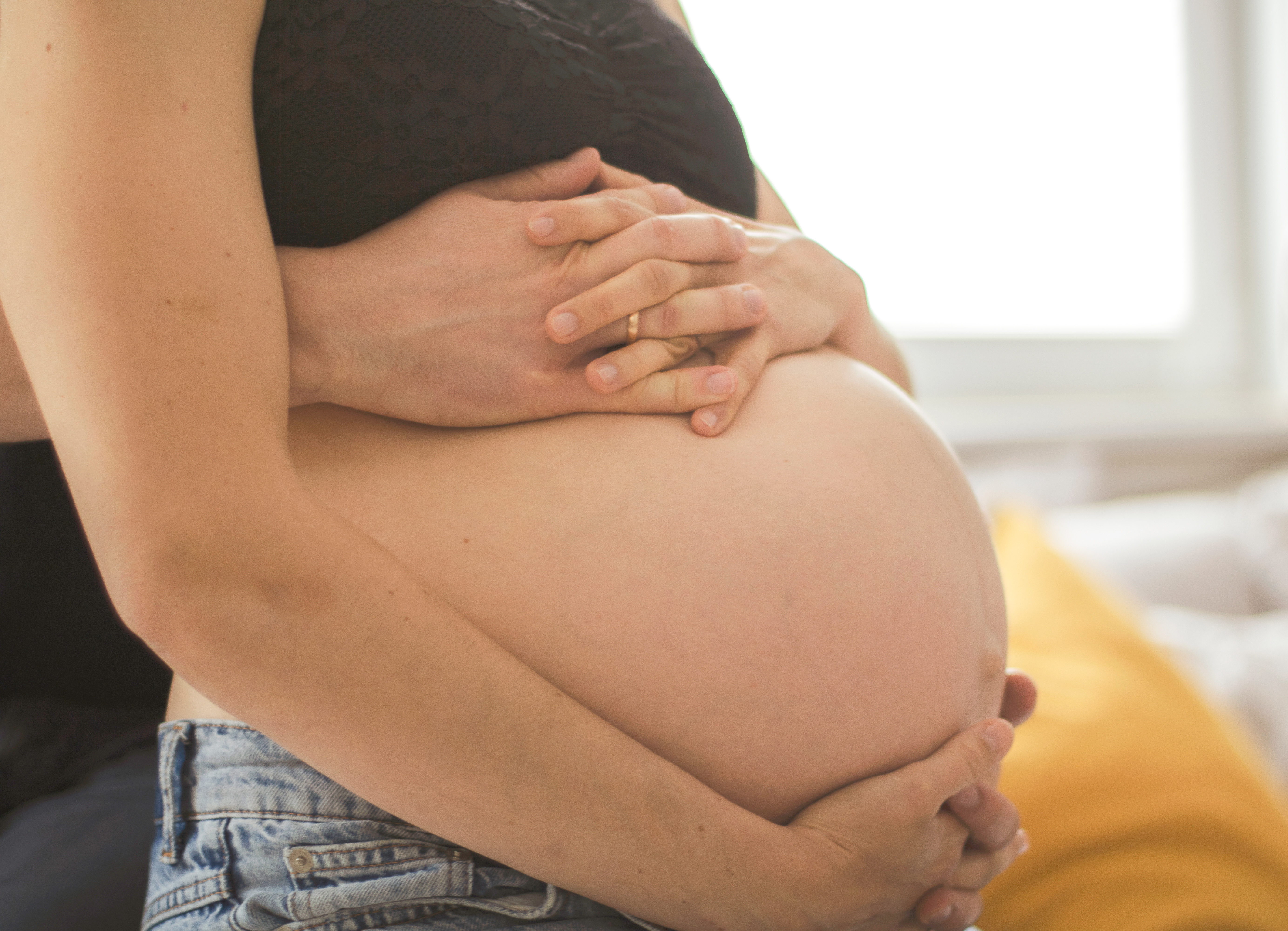 Hände eines Paares ruhen auf einem Babybauch im weichen Fensterlicht.