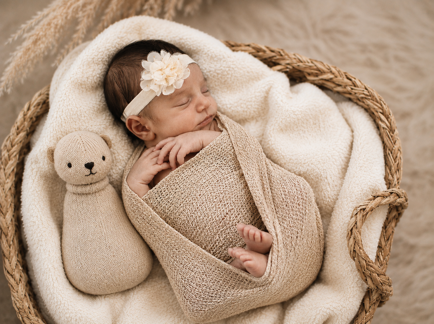 Ein schlafendes Baby mit Blumenhaarband liegt eingehüllt neben einem gestrickten Bären in einem Korb.